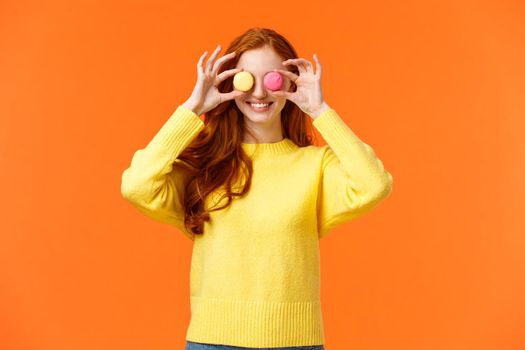Fashion, food and consumer concept. Cheerful silly redhead woman fooling around, playing with dessert, make eyes from macarons and smiling joyfully, standing orange background.