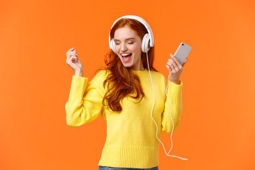 Technology, emotions and gadgets concept. Cheerful good-looking redhead woman dancing with hands up, closed eyes, smiling joyfully, enjoy awesome music sound in headphones, hold smartphone.