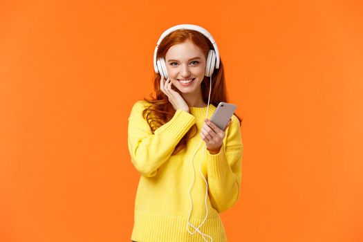 Sassy good-looking redhead female in yellow sweater, listen music white headphones, touch earphones and holding smartphone, listen music, enjoy good earbuds quality, smiling, orange background.