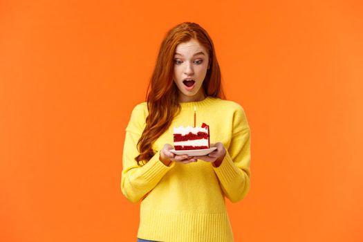 Girl making wish, receive birthday cake with one candle, celebrating b-day, receive surprise feeling amused and grateful, look at dessert with admiration and happy expression, orange background.