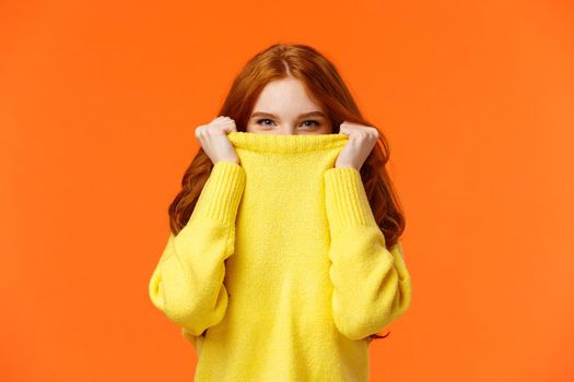Girl hiding from winter cold in her warm soft sweater. Cute redhead woman pulling collar on nose and peeking at camera with smiling eyes, glancing at you happy, orange background.