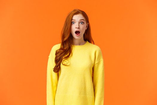 Shocked redhead cute girl drop jaw astounded and speechless, stare camera popped eyes, cant believe, standing impressed and fascinated, losing speech over orange background.