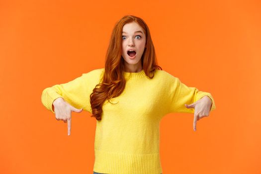 Take look quick. Amazed and fascinated cute ginger girl, redhead student in yellow sweater, gasping open mouth from surprise and wonder, pointing fingers down, look impressed, orange background.
