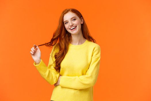 Carefree good-looking, smiling toothy redhead woman, girl with ginger hair laughing as chatting friends, rolling curl on hair, tilt head and smiling joyfully, having conversation, orange background.