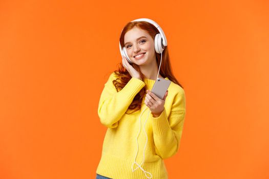 Waist-up portrait cheerful lovely young woman with red hair listen music in headphones, tilt head and smiling satisfied, holding smartphone, pick song for tender calm evening, orange background.