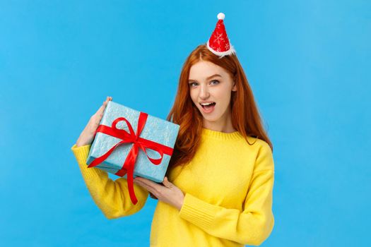 Check-out my gift. Sassy and daring good-looking redhead woman show-off, bragging with wrapped christmas present, looking camera happy and cheeky, wearing cute new year hat, blue background.