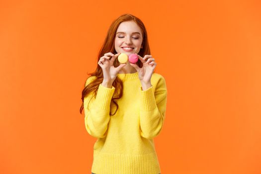 Redhead silly and happy gorgeous girl buy tasty macarons, holding food and looking with desire and affection, like sweets, want eat them, standing orange background satisfied and joyful.