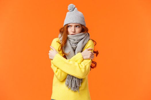 Its freezing cold outside. Cute redhead girl shaking and embracing herself to warm up, waiting someone on street in grey scarf, winter hat during snow and low temprature weather, orange background.