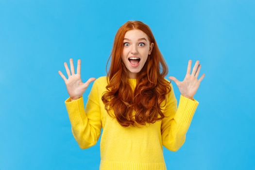 Excited, overwhelmed happy redhead cute girl in yellow sweater, screaming from joy telling about christmas gifts, showing ten fingers, order dozen products, standing joyful blue background.