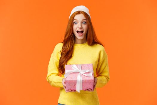 Surprised and excited happy girl unwrapping gifts on christmas eve, new year celebration, smiling amused and thrilled, holding wrapped box present, wearing winter beanie, orange background.
