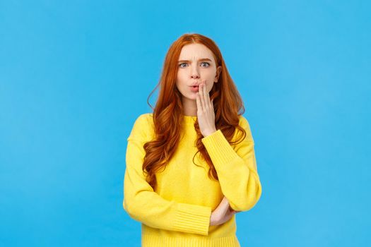 Woman gossiping with colleagues found out shocking revelation. Speechless and concerned young redhead female student heard rumour or bad news, folding lips frowning frustrated, blue background.