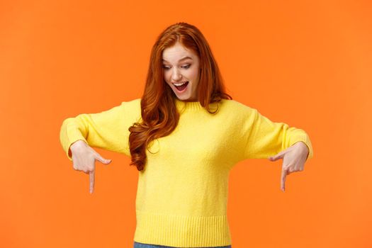 Wondered and excited, amused cute redhead woman in yellow sweater pointing fingers down, look bottom and smiling, check-out product promo, winter holiday event, orange background.