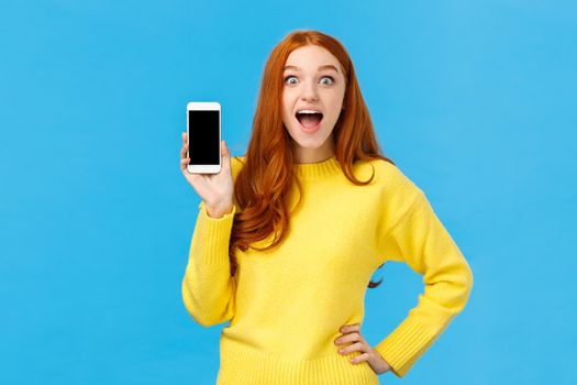 Astonished and impressed, excited redhead female in yellow sweater introduce new app, showing smartphone display, smiling fascinated open mouth amused, look camera, blue background.