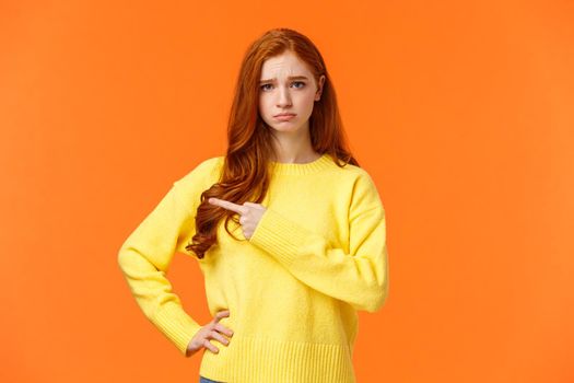 Girl looking upset and unsatisfied. Sad sulky redhead woman pointing left with envy or jealousy cant make it to party, frowning and sulking, showing what she want, standing orange background unhappy.