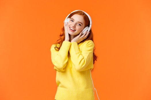 Waist-up portrait cute teenage redhead girl in white headphones, tilt head listen music, touch earphones and smiling camera, singing along favorite song, standing orange background. Copy space