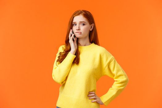 Girl catching anxiety calling someone. Perplexed or nervous cute redhead woman biting lip and looking away as having conversation on mobile phone, standing orange background sad.