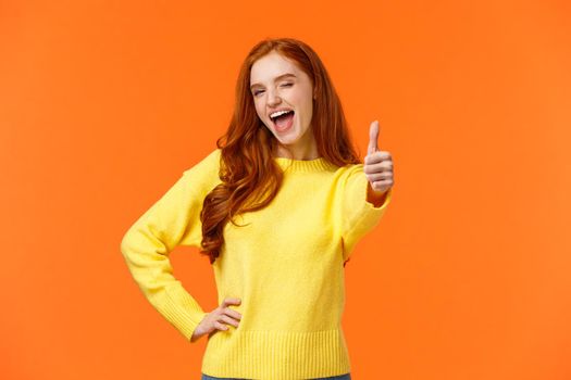 Saying yes. Cheerful redhead woman in yellow sweater giving thumb-up gesture and wink, encouraging keep-up, approving idea, express positive opinion, recommend product or event, orange background.