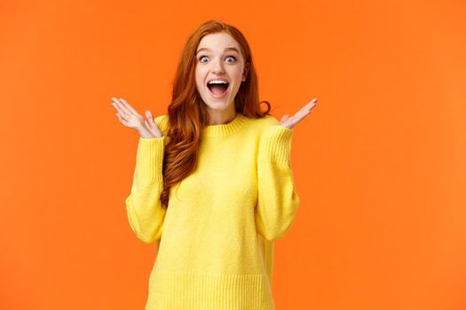 Holidays, surprise and fashion concept. Happy cheerful redhead female customer shopaholic, excited see christmas sales, smiling amused, clap hands from thrill and joy, standing orange background.
