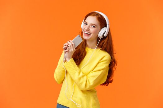 Pretty redhead caucasian woman enjoy listening music, playing karaoke app on smartphone with headphones on, looking camera joyfully, hold mobile phone like microphone, orange background.