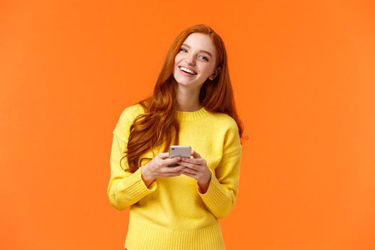 Girl searching gifts for holidays online. Carefree happy redhead female student using smartphone app, laughing joyfully, holding mobile phone, chatting or browsing social networks, orange background.