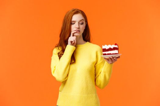 Diet, healthy lifestyle and junk food concept. Cute redhead woman cant resist temptation bite tasty cake, holding dessert on plate and biting lip thinking how many calories in it, orange background.