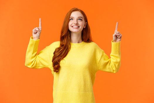 Dreamy happy and upbeat redhead young modern woman gazing up with satisfied smile. Gorgeous girl ginger hair conemplate top advertisement, pointing upwards, standing orange background.