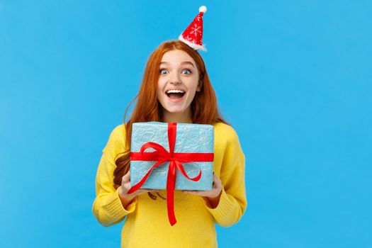 Excited girl love christmas holidays and receiving presents, holding lovely new year gift, smiling joyfully and amused, wearing santa hat as attend party, standing blue background upbeat.