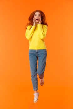 Vertical full-length portrait pretty redhead caucasian girl in casual jeans, yellow sweater, look surprised and excited, holding hands near face impressed, jumping over orange background happy.