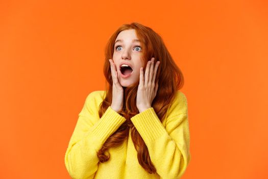Oh no something falling from sky. Shocked and speechless, startled young concerned redhead woman drop jaw, gasping and staring upper left corner concerned, touching face worried, orange background.