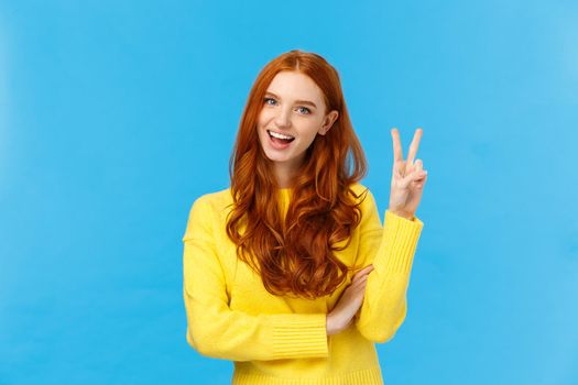 Cheerful outgoing and happy charismatic redhead female in yellow sweater, showing peace sign, number two or twice and smiling, making purchase, order double cheese, blue background.