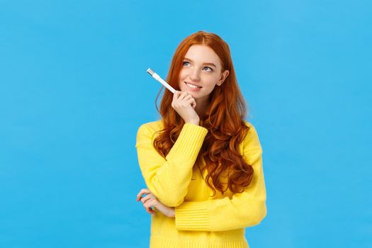 Creative and inspired cute excited redhead woman, journalist or writer creating new ideas, looking upper left corner thoughtful, smiling holding pen as thinking new plot for novel, blue background.