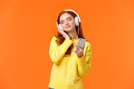 Cute, lovely and tender redhead caucasian female in headphones, touching earphones and close eyes with sensual satisfied smile, listen favorite melody, like song, hold smartphone.