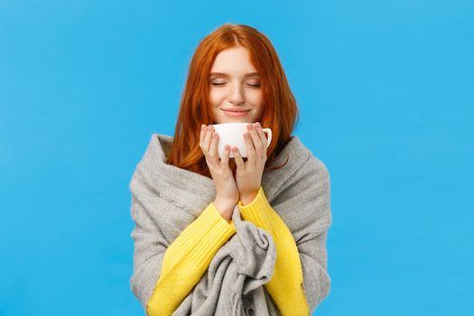 Girl enjoying nice hot cup of coffee smelling flavour with closed eyes and delighted, pleased smile, wrapping herself warm grey scarf, warming during winter low temprature weather, blue background.
