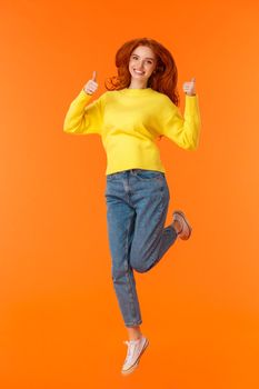 Shopping, fashion and women concept. Vertical full-length positive and cute redhead young modern hipster girl in denim jeans, winter yellow sweater jumping and showing thumbs-up in approval, smiling.