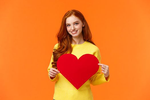 Be my valentine. Lovely gorgeous redhead curly silly girlfriend holding large heart sign and smiling, waiting for valentines day holiday to express love, standing orange background cute.