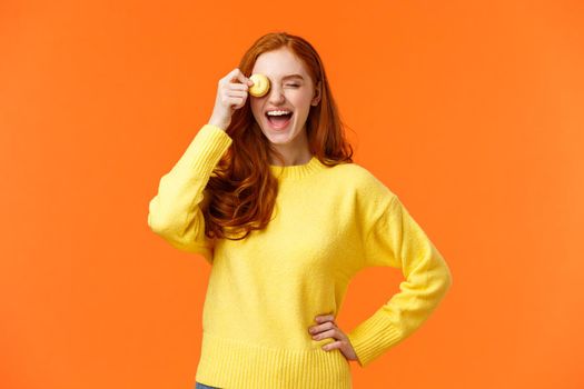 Happy and carefree smiling, laughing lovely redhead girl grinning, holding tasty macaron near eye, fooling around, having fun, eating delicious dessert at favorite cafe, orange background.