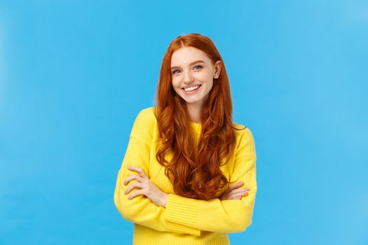 Waist-up shot fashionable pretty hipster girl with red hair, blue eyes, freckles, smiling lovely with cheerful expression, hold hands crossed over chest, confident professional pose, blue background.