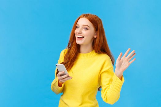 Cute and friendly, outgoing woman with red hair holding smartphone, messaging and waving at someone to turn attention, saying hi, smiling with hello, standing blue background. Copy space