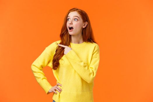 Impressed redhead woman checking-out awesome product sale, stare and pointing left, popping eyes on promo and drop jaw from amazement, surprised, telling about cool advertisiment, orange background.