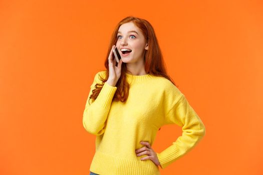 Amused and surprised smiling redhead girl widen eyes and look impressed as hear good news from friend during talking on phone, holding smartphone near ear, have conversation, orange background.