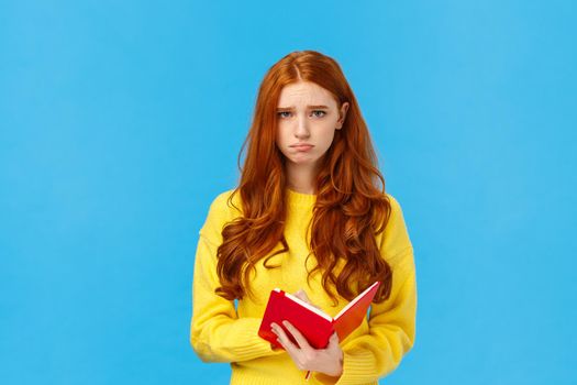 Upset and uneasy, sad cute redhead girl in yellow sweater, sulking frowning and looking camera depressed, holding red notebook, read someones diary, sharing thoughts on paper, distressed.