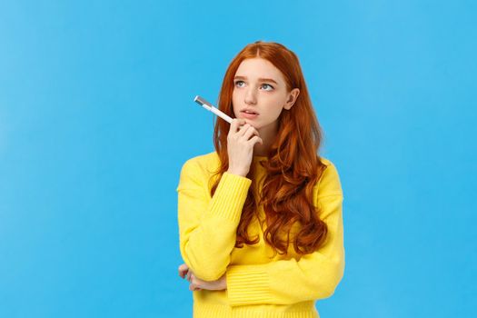 Troubled, hesitant and indecisive redhead, ginger girl with curly hair in yellow sweater, looking unsure upper left corner as pondering, solving exercise in mind, holding pen, searching inspiration.