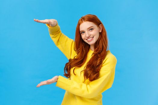 Cute friendly-looking caucasian redhead woman shaping large object with hands, presenting gift, product advertising over blue background, smiling pleasantly, bragging big birthday present.