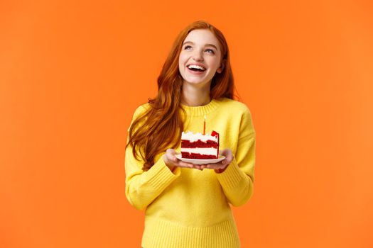 Dreamy happy girl celebrating birthday, having fun, laughing and gazing upper left corner thoughtful, making wish as blowing out candle on delicious, tasty piece b-day cake, orange background.
