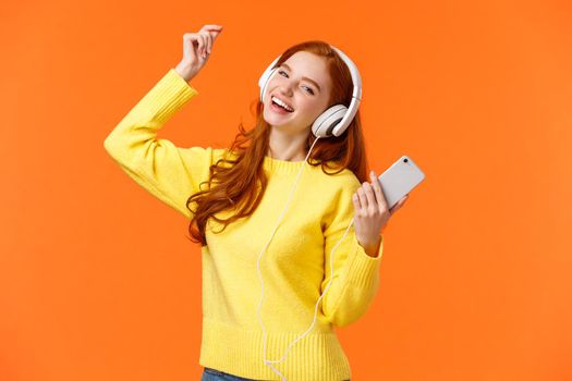 Carefree happy redhead teenage girl receive new headphones for christmas gift, dancing lift hand up while listen favorite music, holding smartphone, put awesome track, orange background.