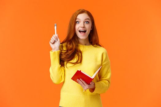 Inspired and dreamy, smart redhead female student got excellent idea for her book, write in notebook plans and to-do list, raising pen in eureka gesture and smiling astonished, orange background.