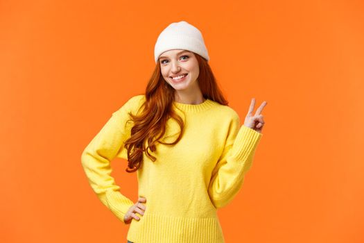 Modern hipster girl playing snowballs with friends during winter holidays, showing peace sign as posing photo near snowman, wearing white beanie and sweater over orange background.