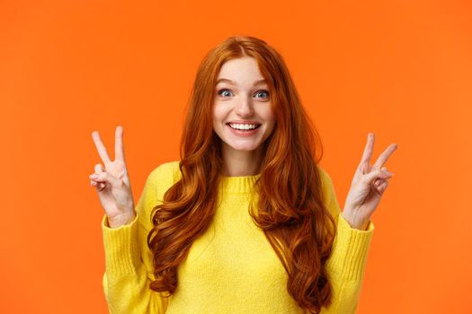 Waist-up portrait lovely and cheerful pretty redhead woman showing peace gestures, smiling camera, travel abroad during winter holidays posing near sightseeing, orange background.