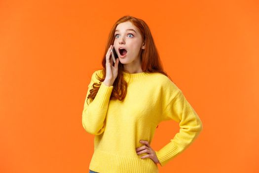 Surprised, startled good-looking redhead female hear amazing news as talking on phone, drop jaw stare left and holding smartphone near ear during conversation, standing orange background.