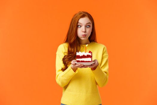 Funny and silly cute redhead b-day girl squinting eyes making goofy expression, pouting as holding birthday cake and blowing-out candle to make wish, having fun at party celebrating, orange wall.
